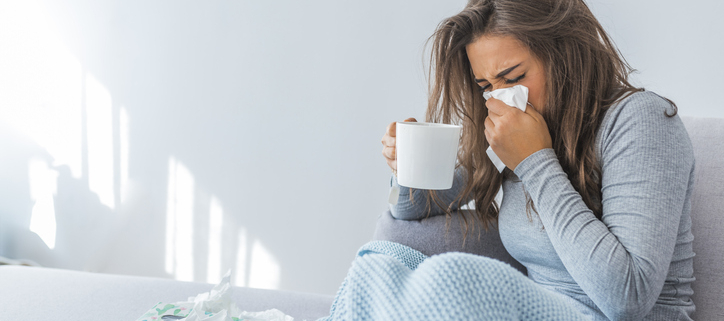 An image of a woman with nasal congestion sitting on a couch wiping her nose with a tissue.